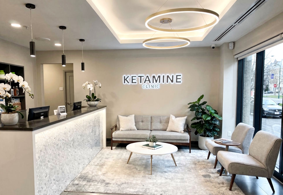 A modern waiting area in a ketamine clinic with a sofa, two chairs, coffee table, potted plants, and reception counter&mdash;perfect for Ketamine Clinic Digital Marketing photos. A "KETAMINE CLINIC" sign adorns the wall and large windows fill the space with natural light. - Ketamine